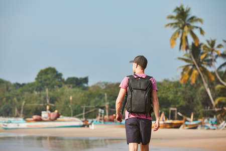Man walking on sand beach against coastline with fishing boats. Rear view of tourist with backpack in Sri Lanka.の写真素材