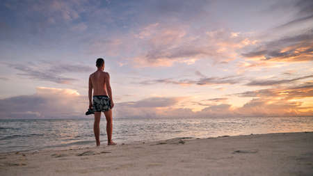 Rear view of young man on idyllic white sand beach. Tourist in swimwear watching colorful sunset in Maldives.の写真素材
