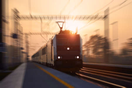 Passenger train fast passing through railroad station. Railway at sunrise with blur effect.