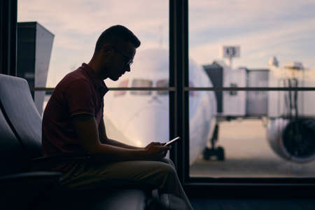 Silhouette of young man waiting while waiting for flight. Traveler using phone in airport terminal.