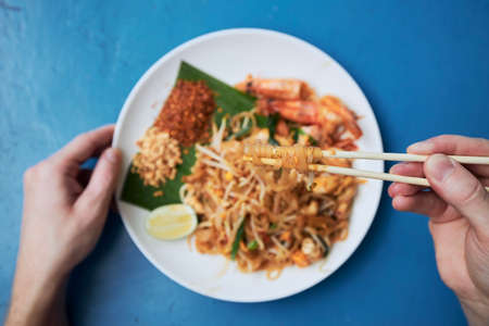 Hand holding chopsticks with meal above plate. Man eating Pad Thai food in street restaurant.の写真素材