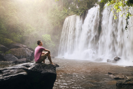 Man sitting on rock in front of high waterfall in mountains in tropical landscape in Camobodia.の写真素材