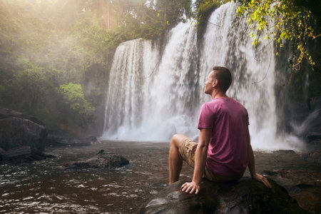 Man resting on stone in front of high waterfall in mountains in tropical landscape in Camobodiaの写真素材