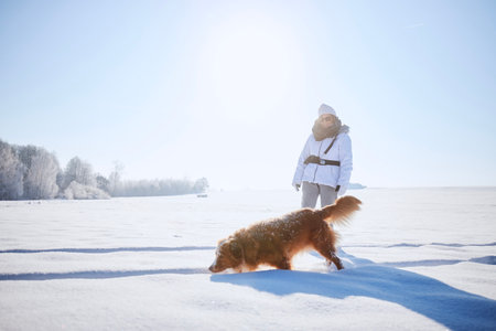 Woman with dog during frosty winter day. Pet owner walking on snowy field with his loyal Nova Scotia Duck Tolling Retriever.の写真素材