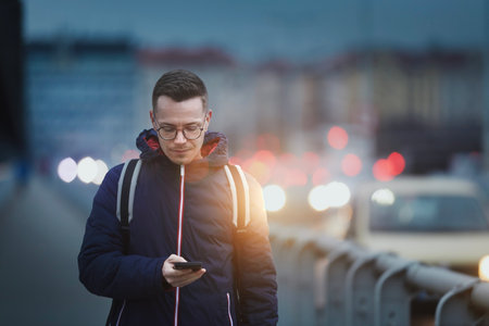 Man looking at phone while walking on busy city street. Urban scene with cars at twilight.の写真素材