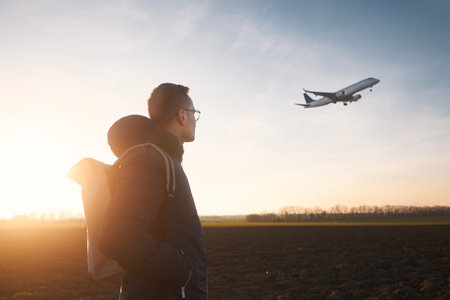 Man with backpack looking up to airplane landing at airport during beautiful sunset.