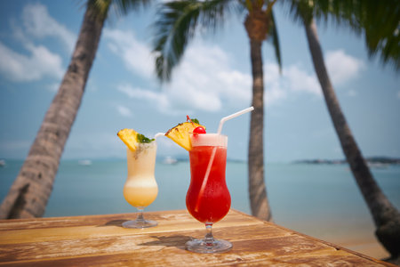 Two cocktails under palm trees on beautiful beach. Singapore Sling and Pina Colada drinks on wooden table.の写真素材