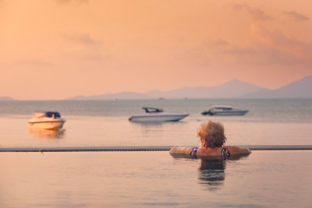 Active senior woman in swimming pool. Rear view of woman enjoying relax and watching sea in sunset.の写真素材
