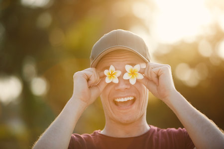 Funny portrait of happy traveler in tropical destination. Man in cap is holding flowers of pumeria in front of his eyes.の写真素材