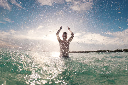 Silhouette of joyful man standing in sea and splashing water towards camera. Sunny day in tropical destination during beach holiday.の写真素材