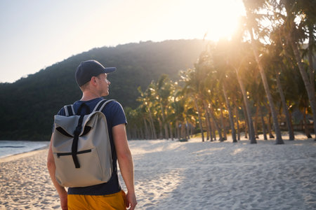 Rear view of tourist with backpack walking on beautiful sand beach with palm trees and looking at sunset.の写真素材
