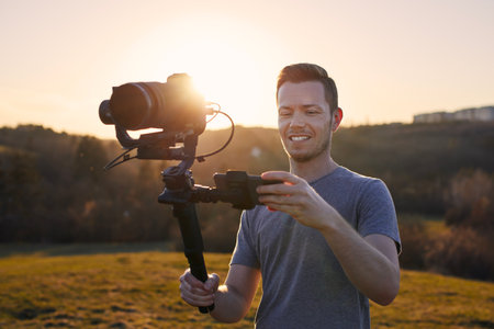 Smiling man while filming with camera and gimbal. Portrait of happy videographer against landscape at beautiful sunset.の写真素材