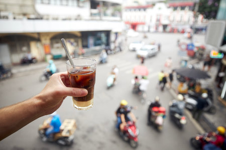 Hand holding of glass of Vietnamese iced coffee with condensed milk. Man drinking coffee above busy...