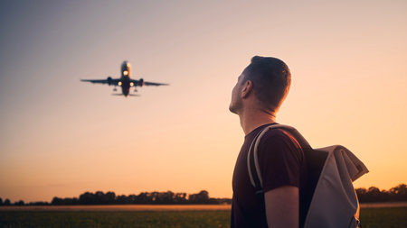 Man with backpack looking up to airplane landing at airport during beautiful summer sunset.の写真素材