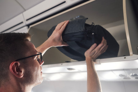Man with backpack is travel by airplane. Passenger putting hand baggage in lockers above seats of plane.の写真素材