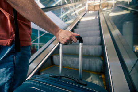 Traveling by airplane. Man standing on escalator in airport terminal. Selective focus on hand holding suitcase. Passenger is ready for travel.の写真素材