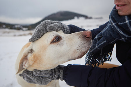 Man dressed in warm jacket, scarf and knitted gloves stroking his dog. Pet owner and labrador retriever against mountain landscape in winter.の写真素材