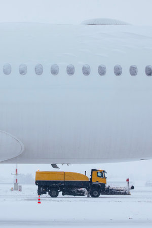 Winter frosty day at airport during heavy snowfall. Airplane covered with snow against snowplows clearing airport runway.の写真素材