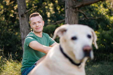 Portrait of man with cute dog sitting in grass under tree. Happy owner of labrador retriever resting in nature on sunny summer day.の写真素材