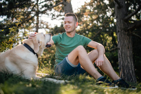 Portrait of man with cute dog sitting in grass under tree. Happy owner of labrador retriever resting in nature on sunny summer day.の写真素材