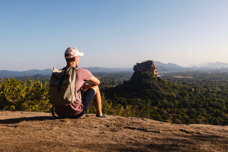 Man with backpack sitting on rock and looking at landscape. Beautiful scenery with Sigiriya rock. Solo traveler in Sri Lanka.の写真素材