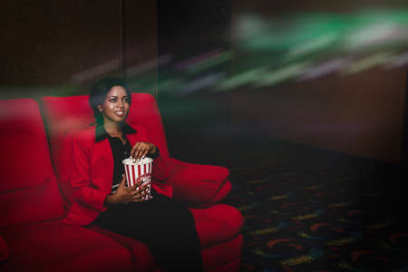 Beautiful African American woman sits watching a movie alone on the red sofa and picks up some canned popcorn to eat as a great time to watch her entertainment.の写真素材