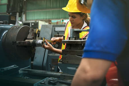 Female technician, engineering department and a colleague working on a lathe, using a vernier caliper to measure the precision steel shaft size, work with professional safety in an industrial factory.の写真素材