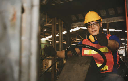 Handsome Asian male worker in the driving division drives a forklift truck to transport goods to an industrial factory ready to perform Thumb up with safety and rigidityの写真素材