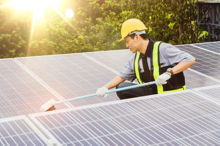 Asian technician is cleaning a blue outdoor solar panel using a brush to scrub away dirt for...
