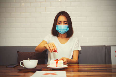 Woman wearing a mask enters a coffee shop, eats cake and hot coffee, sits alone on a separate table according to the Lockdown measure for safetyの写真素材