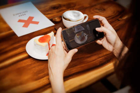 Women like to take pictures of food and snacks before eating in restaurants : Close-up of a female customer hand using a smartphone to take a photo of the cake on the wooden table as a souvenir.