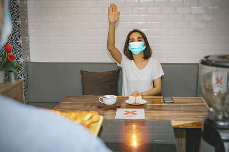 Female customer wearing a mask calls a bisista to serve a coffee shop to order food or check bills (Social distancing concept) : Use in restaurants during the coronavirus (COVID-19) outbreakの写真素材