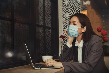 Asian businesswoman wearing a mask, sitting vacantly with depression, by the window of a coffee shop : She was stressed out of work, wanted to be alone and thoughtful.の写真素材