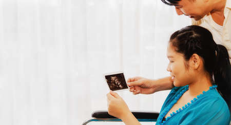 Health-care husband couple, a pregnant wife, in a wheelchair together, look at the ultrasound image of the fetus with a smile, with the expectation of meeting each other at the time of delivery.の写真素材