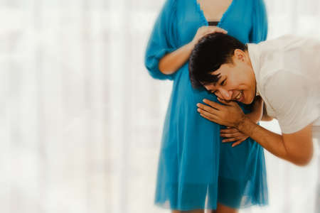 Asian husband is blessed with his near-birth pregnant wife, using their hands and ears to touch the movements of the unborn child with a smile in their home.の写真素材