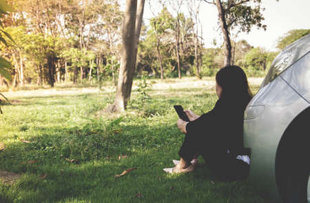 Businesswoman sitting in front of her car in a place full of trees and grasses in the backcountry area,
She is looking for a solution by contacting for help.の写真素材