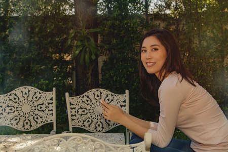 Long-haired Asian woman sitting at an alloy table relaxing in the morning in the front garden, her hobby is to use a cell phone to sit comfortably in her spare time.の写真素材
