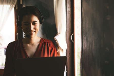 Asian businesswoman wearing a wireless headset working at home with notebook computer, relexing with E-business beside the windowの写真素材