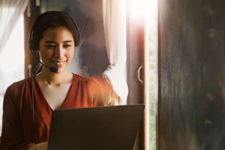 Concept work from home : Asian business woman wearing a wireless headset working at home with notebook computer job freelance, talks to video conference with clients by the window in the morning.の写真素材