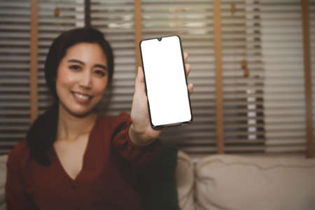 Close up mobile phone : Portrait smiling asian woman showing a white blank screen on her mobile phone while sitting in the house.の写真素材