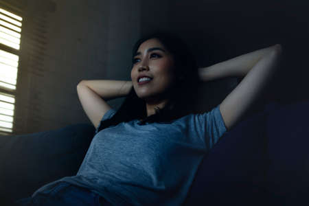 Concept young woman resting at home : beautiful Asian woman sits on a cozy corner sofa in the house by the window of the living room at dusk. She was smiling, carefree, stress-free, and positive.の写真素材