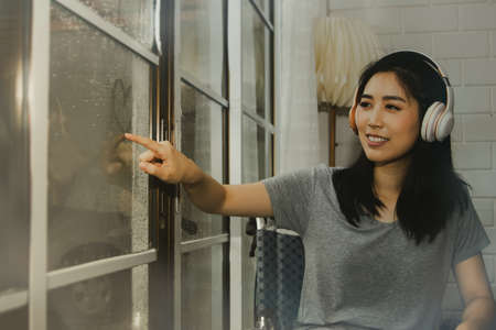 Young, beautiful Asian woman with a lower body disability sits a wheelchair in the house beside the front glass door.She draws a heart shape near the glass with the raindrops on it, happy and relaxedの写真素材