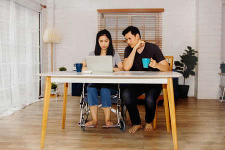 Love concept of a physically disabled couple : Handicapped woman sitting in a wheelchair, working in a living room with her boyfriend, caring, and pampering together willingly.の写真素材