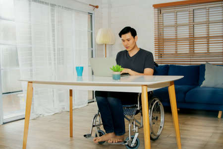 Asian handicapped man in a wheelchair uses his abilities to efficiently work on a laptop computer on a table in the home, despite the fact that his body is sick and ill with paralysis.の写真素材