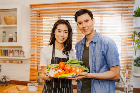 Happy couple in the kitchen : Young Asian couples in the kitchen prepare healthy vegetables and prepare delicious meals together while smiling in their home kitchen.の写真素材