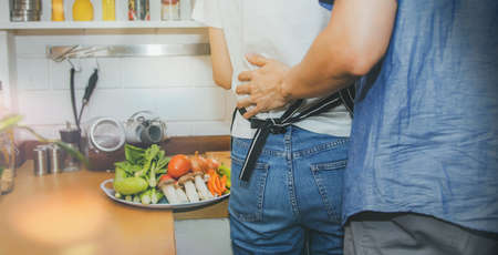 Rear view couple lovingly hugged their waist while preparing for cooking and fresh vegetables in the kitchen.の写真素材