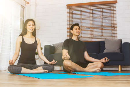 Yoga at home concept : Handsome and beautiful Asian couple meditate and practice yoga, calm their minds with lotus pose on a yoga mat in the living room.の写真素材