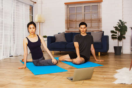 Yoga at home concept : Handsome and beautiful asian couple meditate and practice yoga on their laptop, calm their minds on a yoga mat in the living room.の写真素材