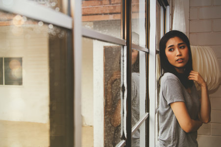 Portrait young asian woman stood near the window, watching the drizzle in loneliness, suffering alone in her heart, thinking of someone to return.の写真素材
