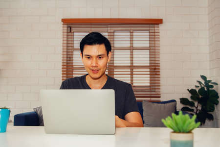Handsome Asian business man wearing a casual T-shirt is working on the internet, social networks, online with a smiling face looking at the screen of his laptop computer : Work from home conceptの写真素材
